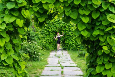 Siyah elbiseli genç, kızıl saçlı bir kadın yeşil parkın arka sokağında mutlu bir şekilde dans ediyor, tırmanan bitkilerle, boş alanla, yaşam sevinci konseptiyle.