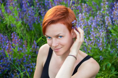 Gülümseyen, kızıl saçlı, kısa saçlı, mavi gözlü genç bir kadının portresi, mavi kır çiçeklerinin (Aconite Dzungarian) arka planına karşı, doğal güzellik kavramı.