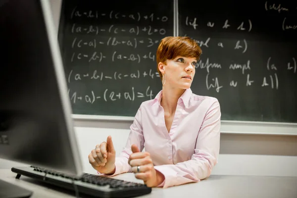 Pretty young college teacher using computer at modern classroom, black chalkboard on background, education concept — Stock Photo, Image