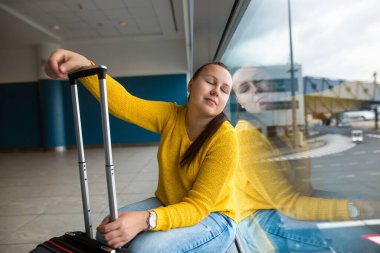Genç esmer kadın bavuluyla havaalanında rötar yapmış uçağını bekliyor, ulaşım konsepti.