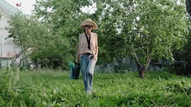 Dziewczyna w słomkowym kapeluszu i dżinsach spacerujących po trawie z konewką — Wideo stockowe