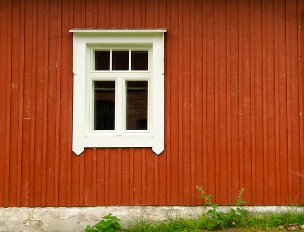 Деревянная стена скандинавского традиционного дома. Стена окрашена в коричневый цвет. В белой рамке есть окно. В нижней части виден фундамент и трава . — стоковое фото