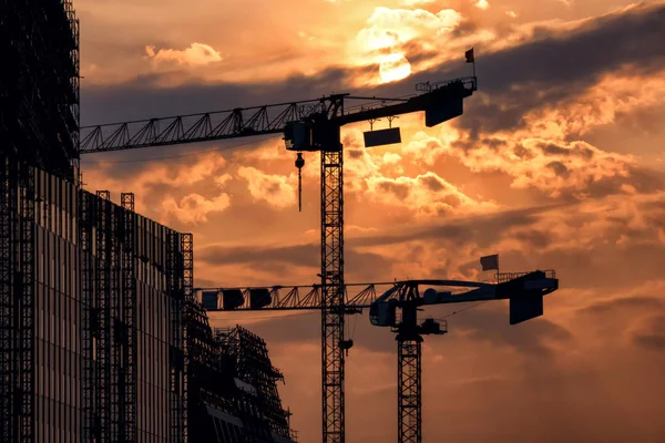 Silhouettes of construction cranes against the evening sky. There are clouds and sun in the sky. On the left there is a construction of a high building under construction. Background. Landscape.