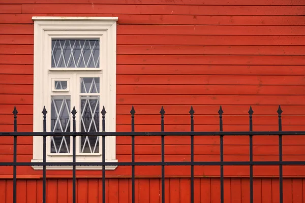 Penceresi olan kırmızı ahşap bir evin manzarası. Ön planda çelik parmaklıklardan bir çit var. Pencerenin beyaz bir ızgarası var. Geçmişi. Doku.
