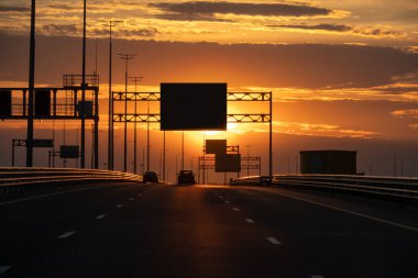 Batıya giden ekspres yolun görüntüsü. Önümüzde güzel bir akşam gökyüzü ve alçak bir güneş var. Büyük karanlık bilgi panoları var. Arkaplan.