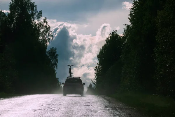 A car with a bicycle on the roof drives on a wet road. There is a tall forest at the edges. In the distance you can see the sky with clouds.