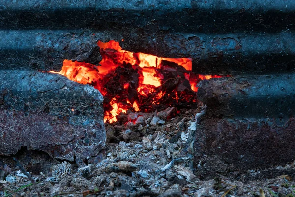 Eski bir çöp yakma varilinde bir delik. Metal eskimiş ve paslanmış. Arka planda yanan kömürler görülebilir. Geçmişi. Doku.