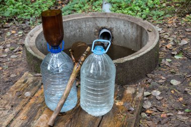 Round concrete well by a spring in the forest. In the foreground are two transparent bottles. One of them is filled with water. There is a primitive scoop and funnel from scrap materials. Background. clipart