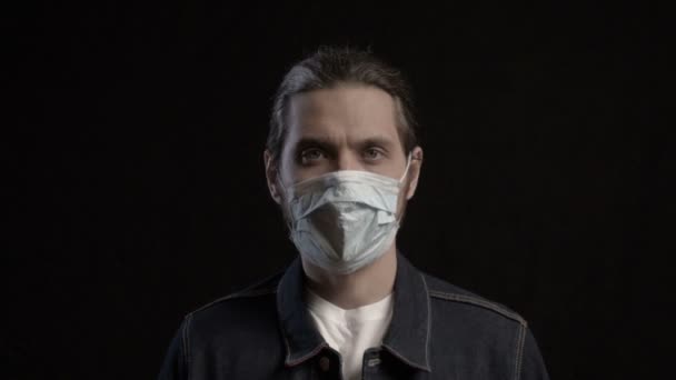 Een volwassen man met een medisch gezichtsmasker, kijkend naar een camera op zwarte achtergrond. Corona virus, Covid 19. Coronaviruspandemie. — Stockvideo