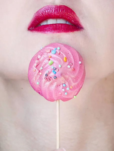 Closeup view of sexy sensual bright female lips with pink lipgloss on mouth at delicious round colorful lollipop candy on stick, meringue