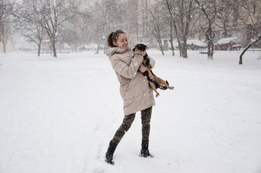 Oldukça sevimli genç kız ya da seksi kadın bej kışın gülümseyen ceket ve askeri kot pantolon doğal beyaz zemin üzerine karlı soğuk kış kar ve kar taneleri ile siyah köpek ya da köpek yavrusu evde beslenen hayvan duruyor kamuflaj