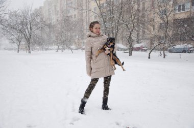 Oldukça sevimli genç kız ya da seksi kadın bej kışın gülümseyen ceket ve askeri kot pantolon doğal beyaz zemin üzerine karlı soğuk kış kar ve kar taneleri ile siyah köpek ya da köpek yavrusu evde beslenen hayvan duruyor kamuflaj