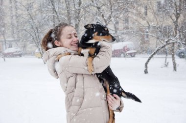 Oldukça sevimli genç kız ya da doğal beyaz zemin üzerine soğuk kış kar ve kar taneleri ile siyah köpek ya da köpek yavrusu evde beslenen hayvan duruyor bej kış ceket karlı seksi kadın gülümseyerek