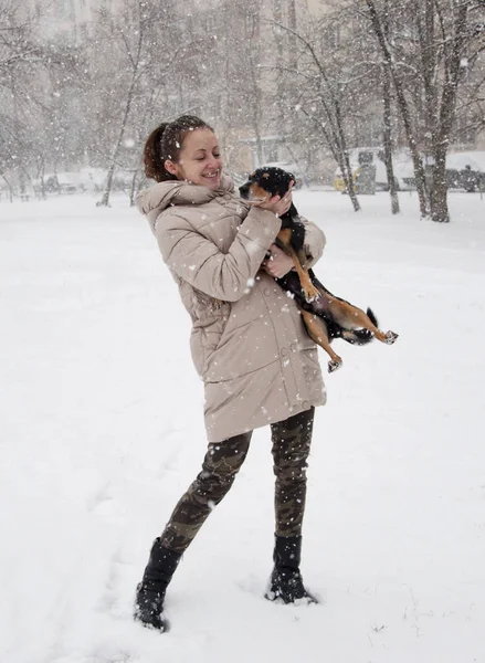漂亮可爱的年轻微笑的女孩或性感的女人在米色冬衣和伪装军事牛仔裤雪冷冬天在自然白色背景站立在雪和雪花与黑狗或宠物小狗 — 图库照片