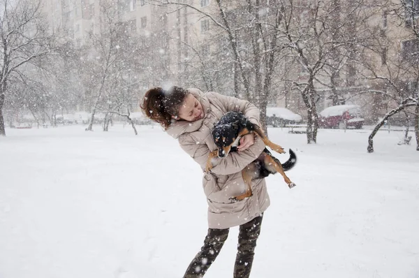 ベージュ冬の女の子やセクシーな女性を笑顔かわいい若いコートし 自然の白い背景の雪の降る寒い冬は雪と黒犬か子犬ペットと雪に立つ軍のジーンズを偽装 — ストック写真