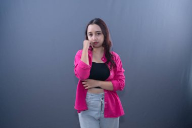 Pembe gömlekli ve kotlu genç bir kadın, düşünceli görünüyor, gri bir arka plana karşı poz veriyor.