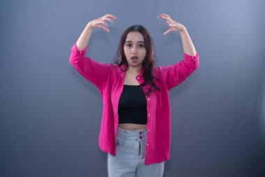 Pembe gömlekli ve siyah ekili bluzlu genç bir kadın, elleri havada ve ağzı açık bir şekilde şok veya şaşkınlık yaşıyor.