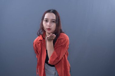 Turuncu tişört giyen genç bir kadın, tarafsız bir ifadeyle izleyiciye öpücük yolluyor.