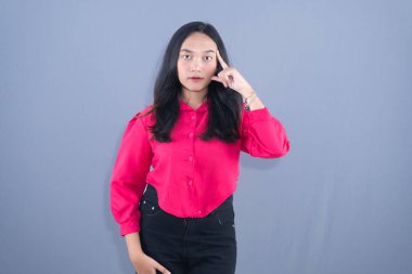 Kırmızı gömlekli ve siyah pantolonlu genç bir Asyalı kadın düşünceli bir jest yapıyor..