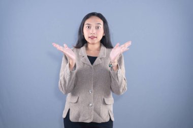 Genç bir Asyalı kadın omuz silkiyor, şaşırmış / kafası karışmış görünüyor gri bir arka plana karşı elleri açık