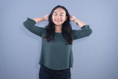 Gözleri kapalı genç bir kadın, sakin bir şekilde gülümsüyor, elleri başının arkasında, düz bir arkaplanda.