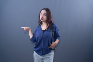 A young Asian woman in a blue cardigan and light pants, pointing left with a surprised expression