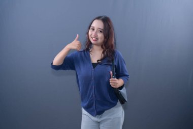 Young Asian woman in blue cardigan, smiling, giving thumbs-up, holding bag, grey background
