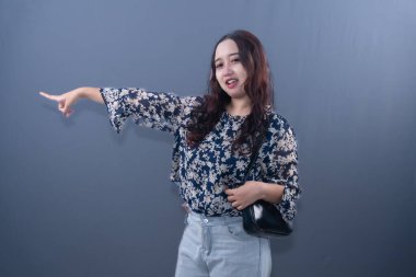 A young Asian woman in floral top, light jeans, black bag, pointing left, smiling on grey background