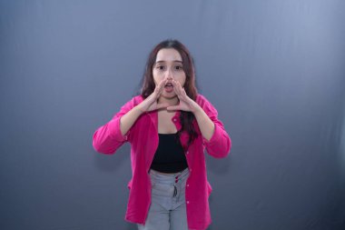 Young woman in pink shirt, hands cupped to mouth, shouting or calling out, on grey background