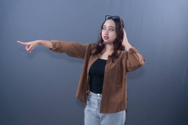 A young Asian woman in brown shirt, black top, and jeans, pointing left, looking surprised