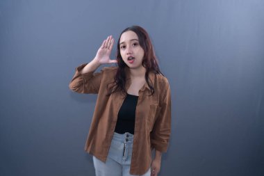 Young Asian woman in brown shirt, black top, and jeans, hand to ear, listening intently