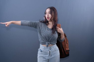 Young Asian woman in grey shirt & jeans, brown backpack, pointing left with surprised expression