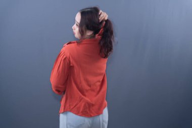 A young Asian woman in an orange top, back to camera, hand in hair, on a grey background