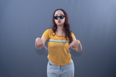 Young Asian woman in yellow striped shirt & sunglasses, pouting, pointing down