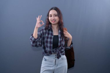 A young Asian woman in plaid shirt & jeans, smiling, making 