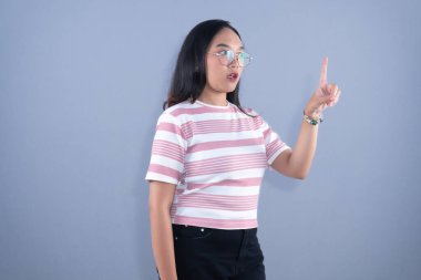 Young Asian woman in striped shirt and glasses, pointing finger upwards, serious expression