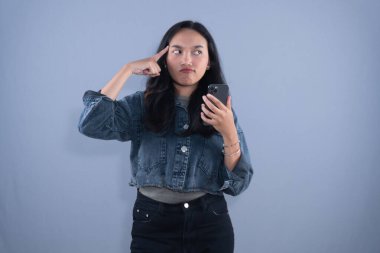 Young Asian woman in denim jacket, holding phone, finger on temple, looking puzzled, grey background