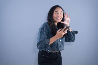 A young Asian woman in a denim jacket, holding a phone, cupping her ear to listen intently