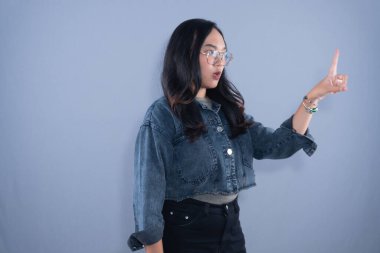 A young Asian woman in a denim jacket and glasses, pointing upwards with a surprised expression