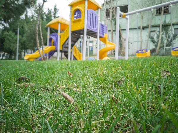 Zelená tráva zavře obraz s pozadím s dětským hřištěm pro děti v parku zelených stromů. — Stock fotografie