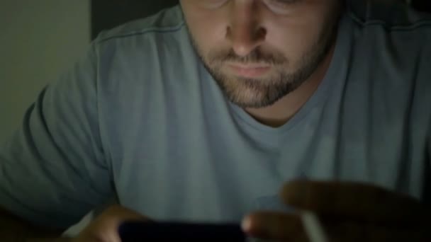 Ein junger Mann hält ein Smartphone in der Hand und raucht in einem dunklen Raum. der bärtige Typ lächelt und lacht. — Stockvideo