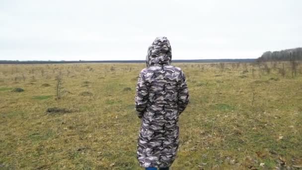 Eine einsame Frau in einer langen Tarnjacke geht an einem großen Feld entlang. Spaziergang an einem ungewöhnlichen Ort. — Stockvideo