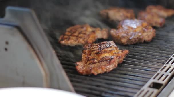 Os bifes de carne são cozinhados em uma grelha a gás. Pratos de carne saborosos ao ar livre. O fumo vem da comida. Cook vira a carne com pinças de metal . — Vídeo de Stock