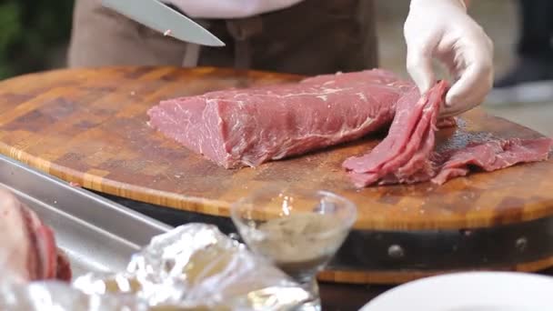 Cook in witte handschoenen afsnijdt, kleine stukjes steaks van een groot stuk van rauw vlees. Varkenshaasje van de kwaliteit voor de bereiding van heerlijke gerechten. Een scherp mes en een houten plank — Stockvideo