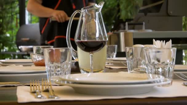 Стол в ресторане сервируется для встречи гостей. Бокал красного вина сливается с кувшином, который стоит позади. Повара на заднем плане готовят что-то на гриле . — стоковое видео