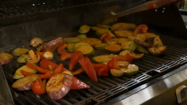 Cook zamienia pomocą kleszcze różnych warzyw, takich jak bakłażan, papryka, cukinia i grzyby. Zdrowe jedzenie jest gotowane na grill. — Wideo stockowe