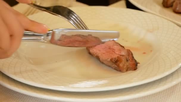 Las manos femeninas con un cuchillo y un tenedor cortan un trozo de filete de carne tostado medio en el plato. El plato se refleja en los cubiertos de metal. Degustación de alta cocina . — Vídeo de stock