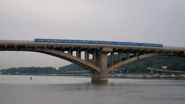 O trem viaja sobre a ponte contra o céu e a floresta. O metro saiu do lado de fora. Transporte público em movimento. Carros atravessam a ponte sobre o rio . — Vídeo de Stock