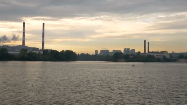 El avión está aterrizando en el fondo de chimeneas industriales y puesta de sol. La vista desde el barco que flota en el agua en la ciudad de la tarde y el cielo . — Vídeo de stock
