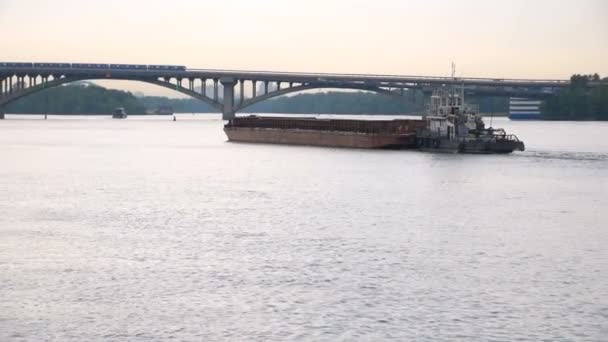 Um pequeno navio reboca uma barcaça com uma carga no rio na direção da ponte . — Vídeo de Stock